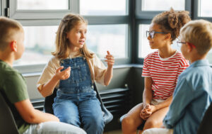 Four children sit in a circle indoors, talking and listening. A girl in dungarees gestures as she speaks about her experiences with epilepsy, while the other three children, two boys and a girl, focus on her. Large windows are in the background.