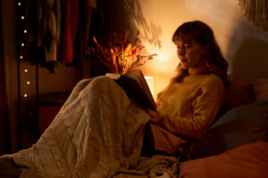 Young adult Caucasian woman reading book in bed under blanket, promoting mental wellbeing and self care. Image suitable for lifestyle blogs and leisure industry marketing, medium shot